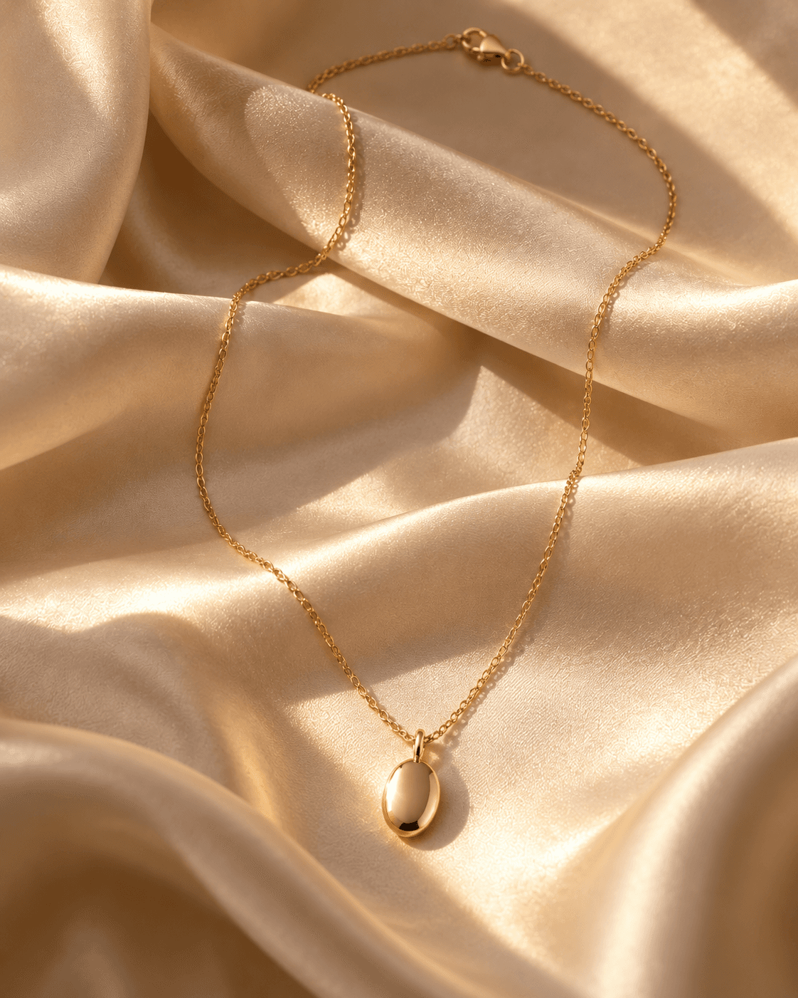 Gold oval pendant necklace draped across folds of champagne satin in warm light