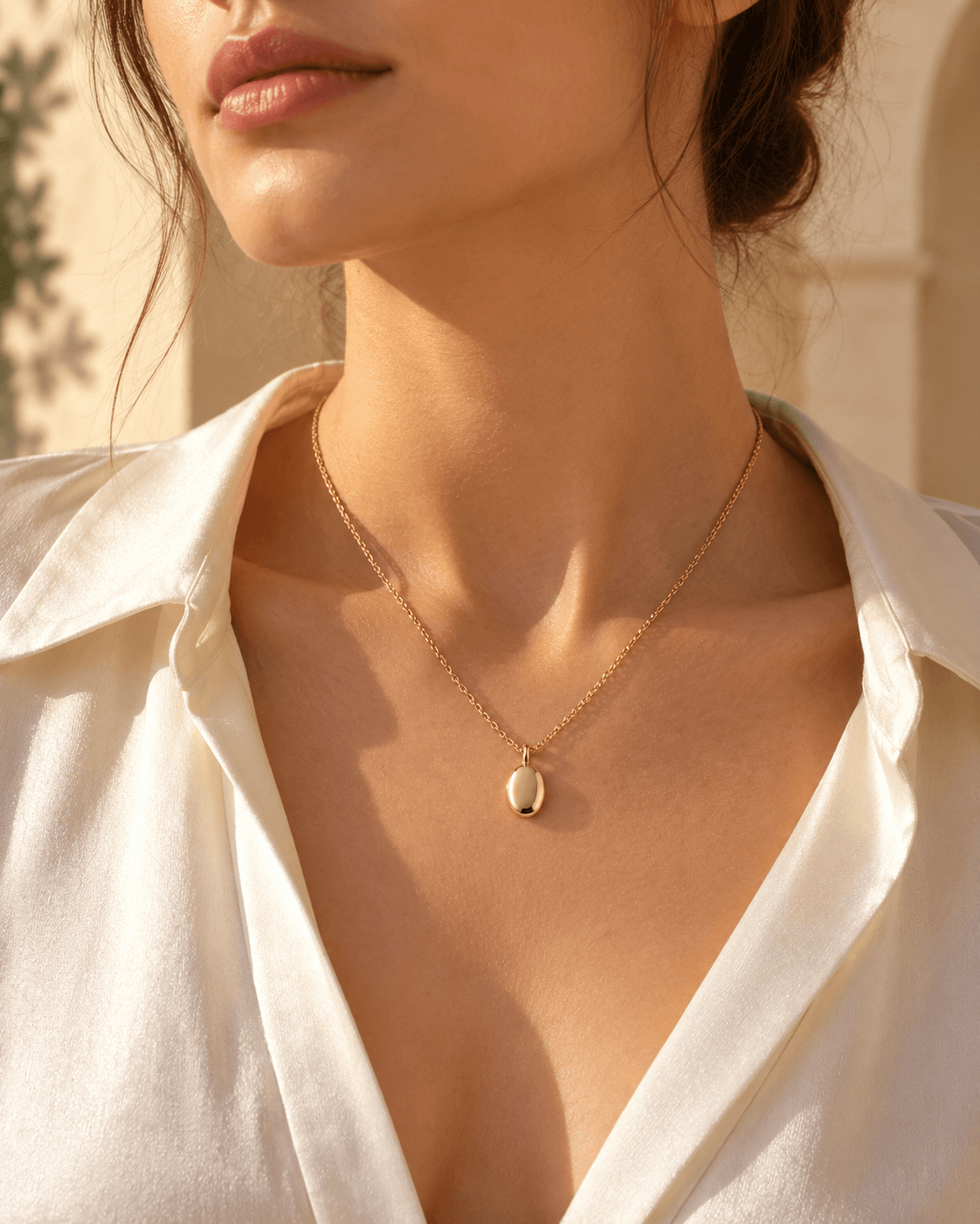 A model wearing the gold pendant necklace with an open white silk shirt, in warm sunlight