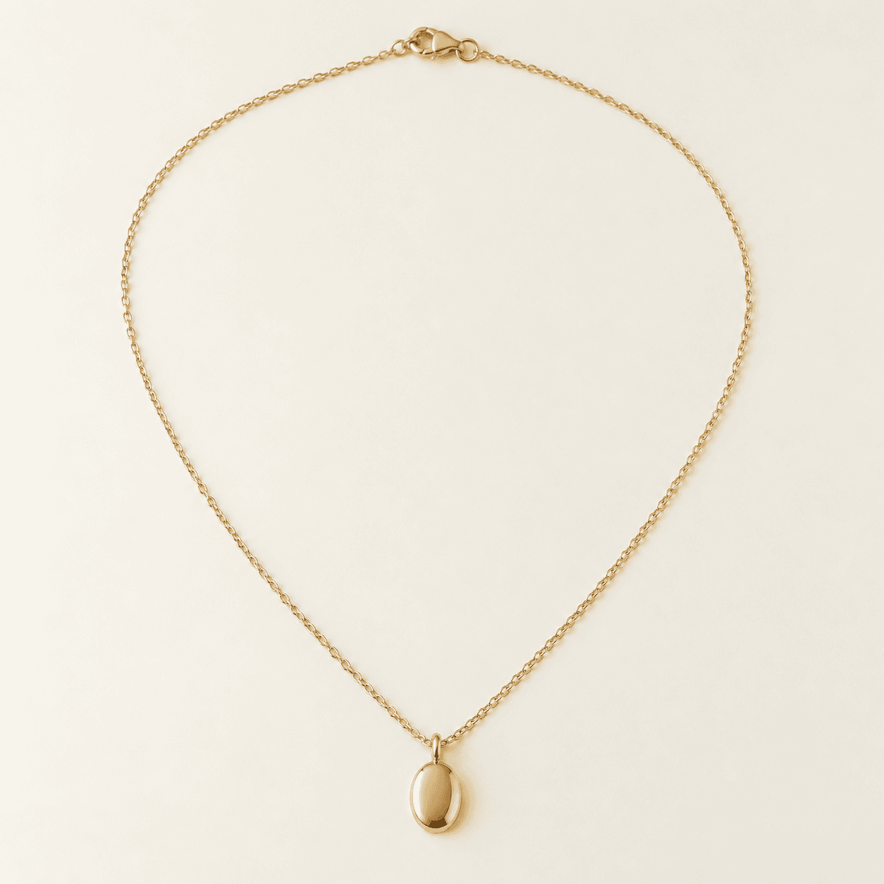 Gold oval pendant necklace on a fine cable chain, laid flat on a plain cream background